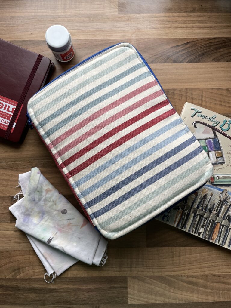 Photo of a fabric case with faded red, white and blue stripes surrounded by sketchbooks, a glue stick and a rag to wipe paint brushes on 