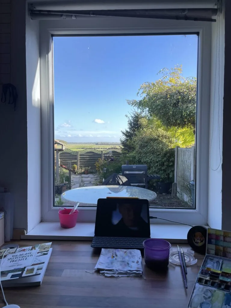 View through a rectangular window out into a garden full of green plants and flowers, overlooking a wide open farmer's field. On the desk is a selection of paint brushes, paints, an iPad and a sketchbook.