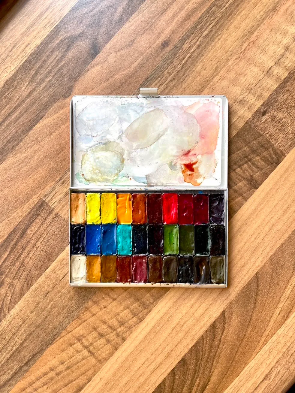Photograph of an open watercolour palette showing 30 rectangular shallow pans of paint, sat on a walnut wood worktop.