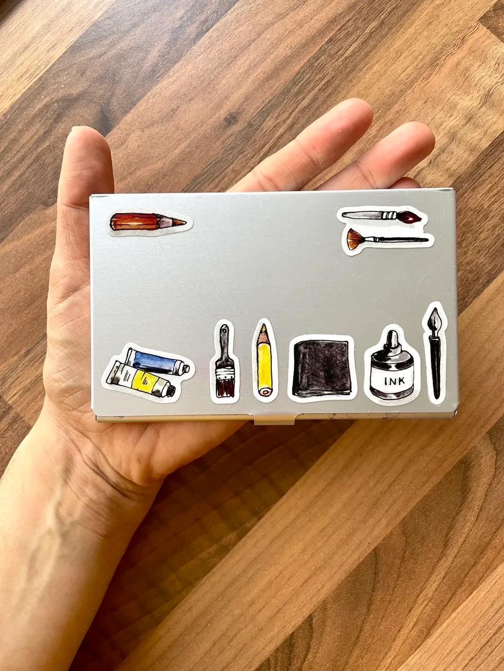 Photo of a closed silver ArtToolKit Folio palette with Samantha Dion Baker stickers of miniature art supplies on.
