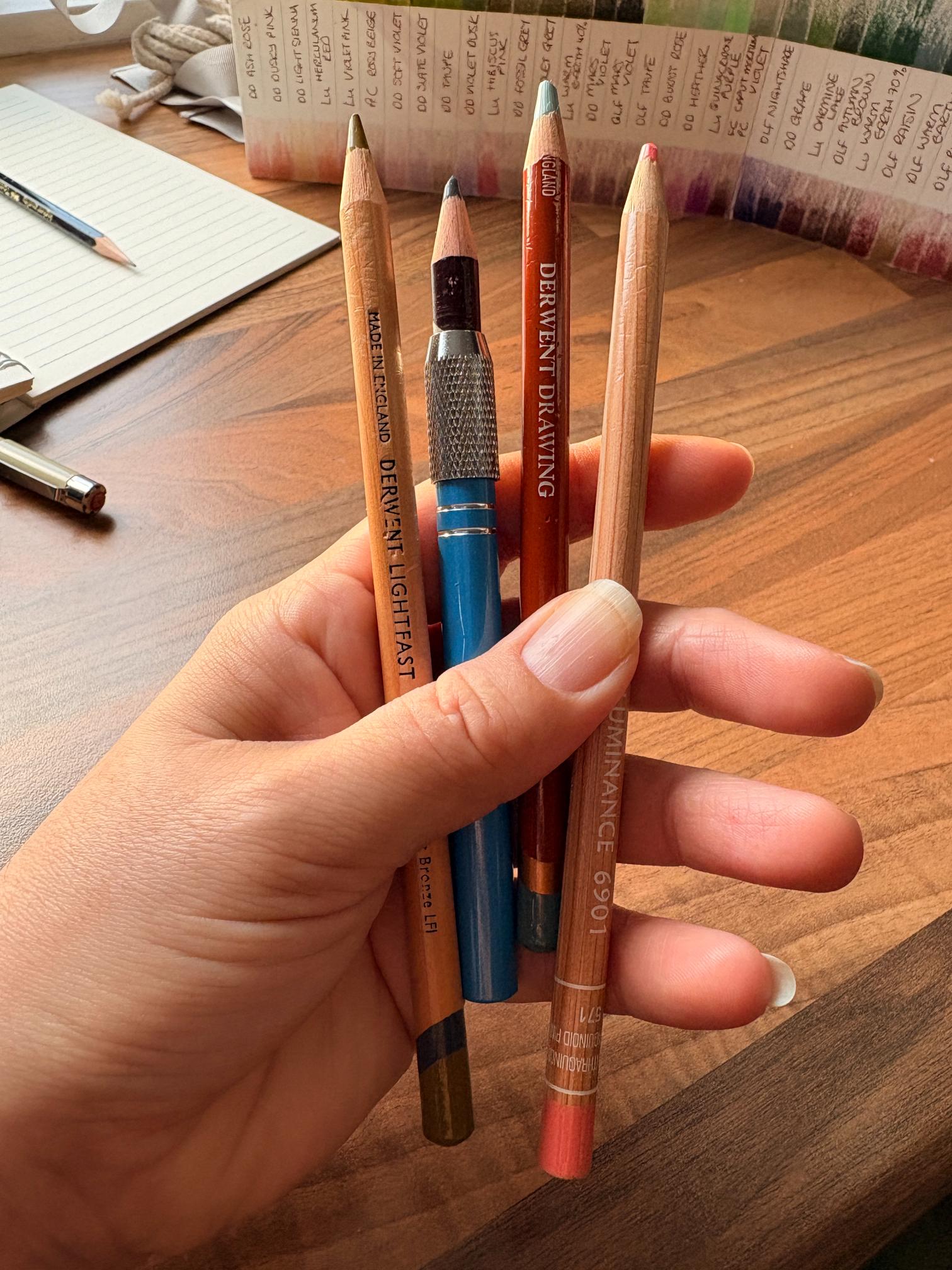 My hand holding a selection of coloured pencils in various brands